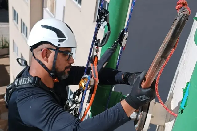 Profissional com capacete trabalhando suspenso por cordas na fachada de um prédio