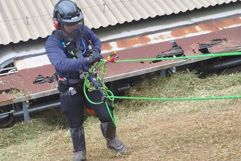 Profissional com capacete e protetor auricular operando cordas de segurança ao lado de uma estrutura industrial